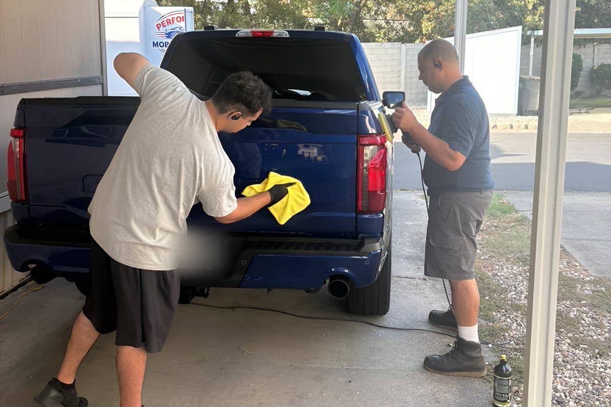 Two people working at the tailgate of a blue Ford F-150, one wiping with a microfiber towel